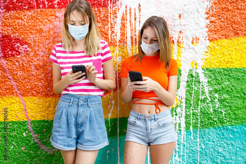 Fashionable female friends in shorts using smart phones against graffiti wall during pandemic photo