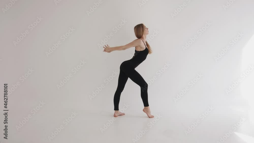 A slender young girl in a black top and leggings is engaged in stretching in a white studio and makes smooth body movements