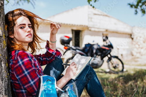 Portrait of redheaded motorcyclist leaning against tree trunk taking notes, Andalusia, Spain