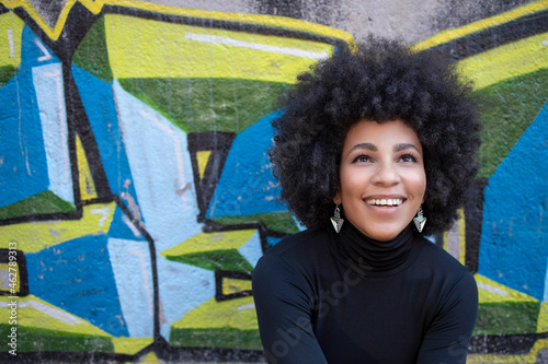 Happy afro young woman looking up while sitting against graffiti wall photo