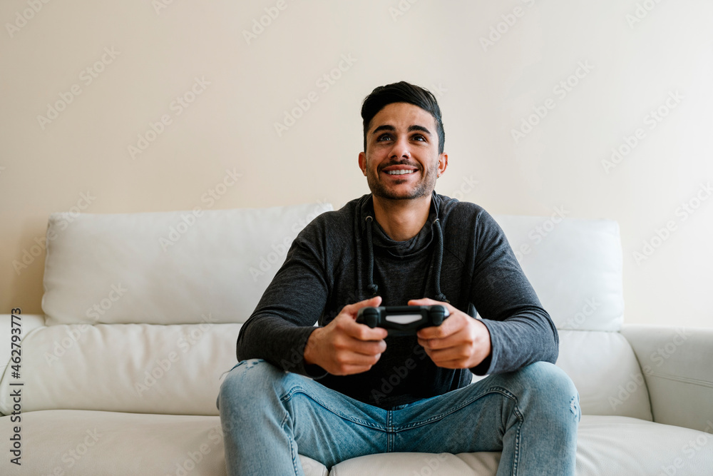 Smiling young man sitting on sofa while playing video game at home Smiling young man sitting on sofa while playing video game at home