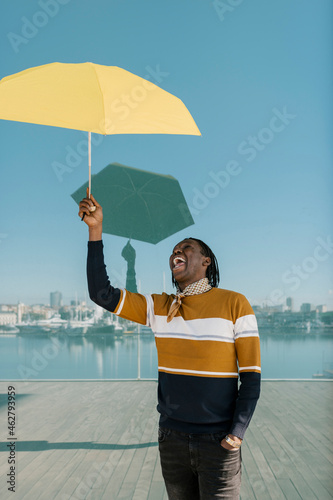 Smiling man with hand in pocket holding yellow umbrella while standing against glass wall photo