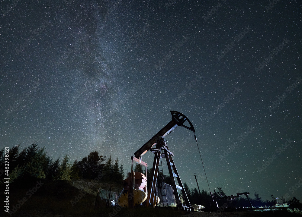 Pumpjack against starry sky with Milky way in natural mountains environment for mining gasoline, pipeline system extract crude oil petroleum and water emulsion at each stroke during night time - obrazy, fototapety, plakaty 