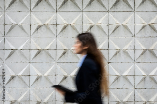 Blurred young businesswoman holding tablet passing concrete wall