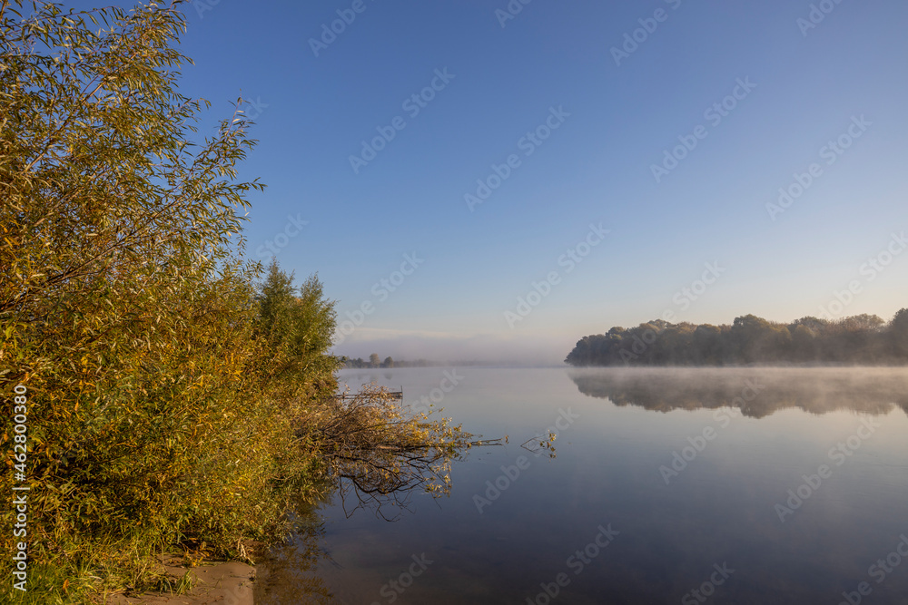 Autumn landscape in the early morning on the river. Misty water surface. Yellow leaves on trees and bushes are illuminated by the rays of the rising sun. Dawn on a cold autumn morning.