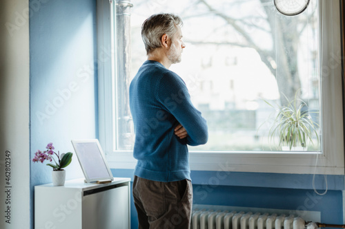 Pensive man standing in living room looking out of window photo