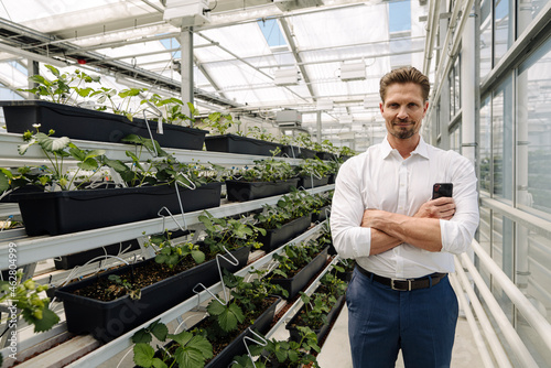 Confident male entrepreneur with arms crossed standing by plants in greenhouse photo