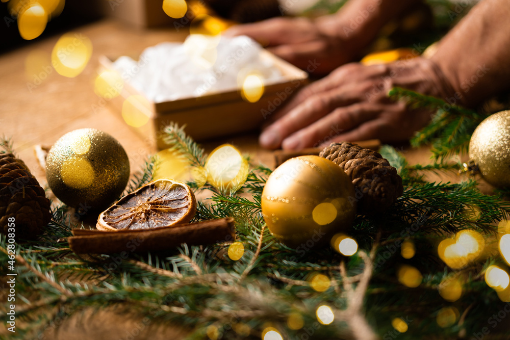 Male hands make a Christmas and New Year's gift on a wooden background branches of a Christmas tree, gold decorations, cinnamon sticks and orange. Christmas composition.