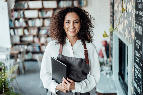 Smiling woman wearing apron holding book while standing at cafe photo