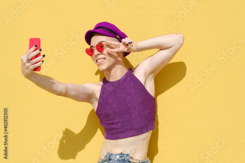 Non-binary young person taking selfies with a mobile phone while standing against a yellow wall.