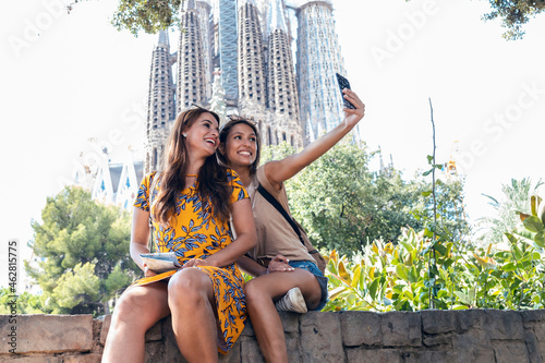 Smiling friends taking selfie while sitting on retaining wall at Sagrada Familia park, Barcelona, Catalonia, Spain photo