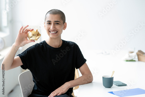 Portrait of a smiling woman holding up golden piggy bank at desk in offce photo