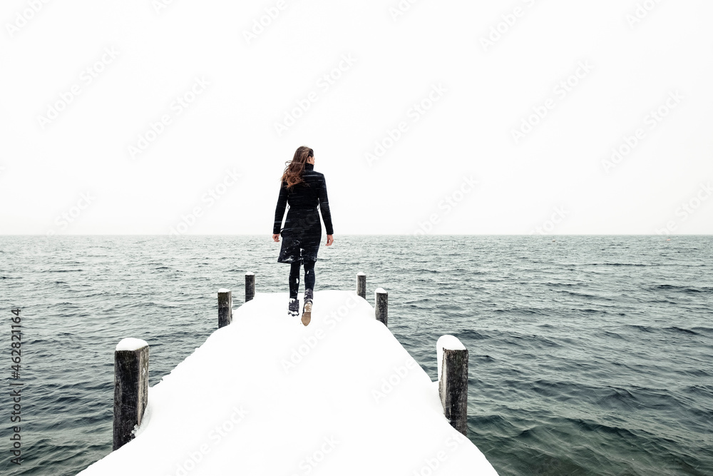 Back view of young woman walking on snow-covered jetty at Lake Starnberg, Germany Back view of young woman walking on snow-covered jetty at Lake Starnberg, Germany