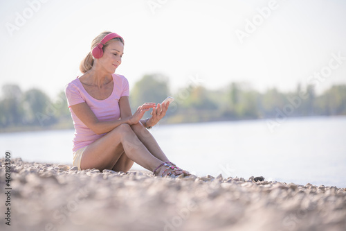 Mature woman relaxing at the riverside listening to music with headphones and using cell phone