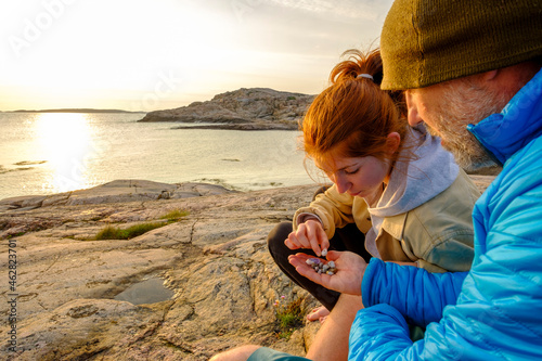 Sweden, Vastra Gotaland County, Grebbestad, Man and teenage girl collecting pebbles on rocky shore of Tjurpannans Nature Preserve at sunset photo