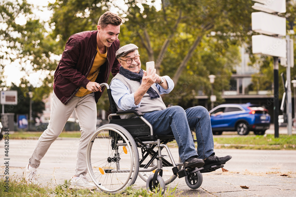 Smiling young man pushing happy senior man with smartphone in wheelchair Smiling young man pushing happy senior man with smartphone in wheelchair