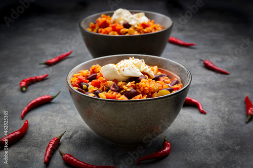 Red chili peppers and two bowls of vegetarian chili with red lentils, kidney beans, tomatoes, carrots, celery and sour cream photo