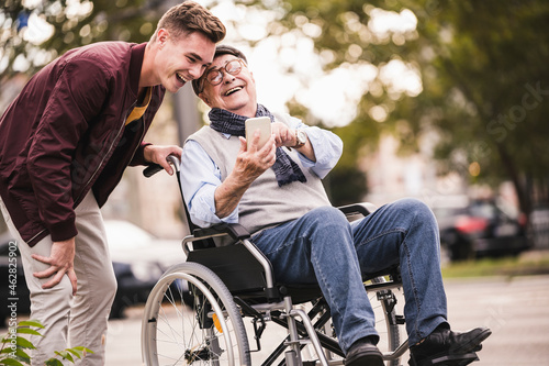Laughing senior man in wheelchair and his adult grandson looking together at smartphone having fun photo