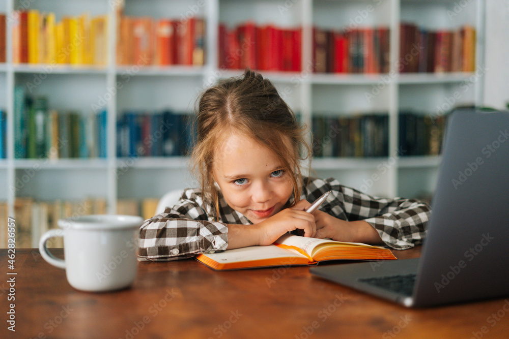Close-up portrait of adorable cheerful pupil child school girl lying at table with laptop and paper notebook, looking at camera. Cute smiling schoolgirl e-learning online using computer