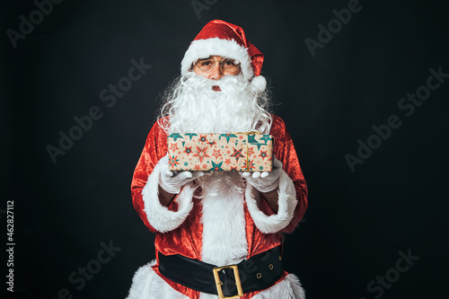 Man dressed as Santa Claus holding a parcel wrapped with wrapping paper, on black background. Christmas concept, Santa Claus, gifts, celebration.