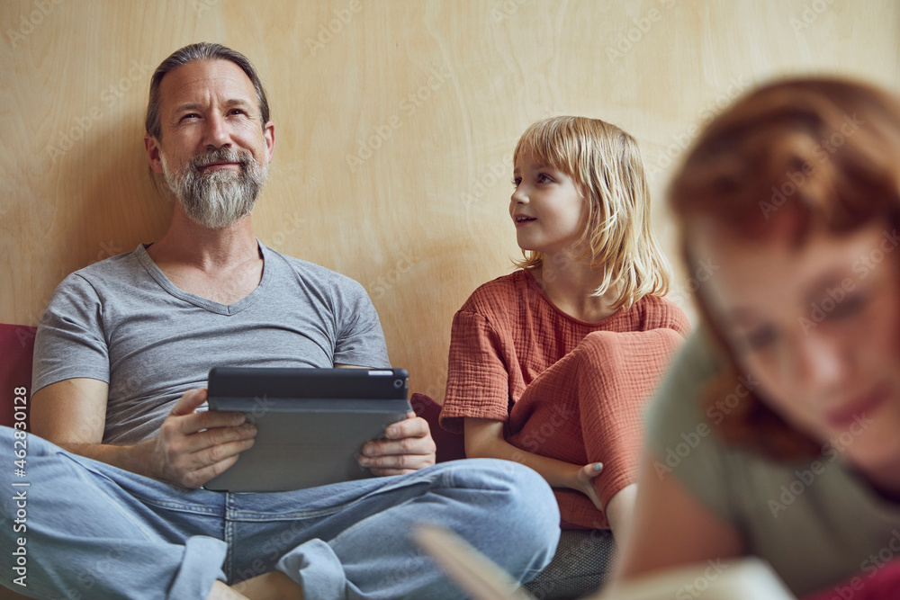 Father and daughter with digital tablet looking away while sitting against wall at home Father and daughter with digital tablet looking away while sitting against wall at home