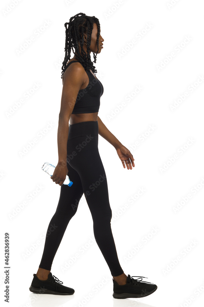 African american woman in black sports clothes is walking with bottle of water. Full length, side view, isolated. African american woman in black sports clothes is walking with bottle of water. Full length, side view, isolated.