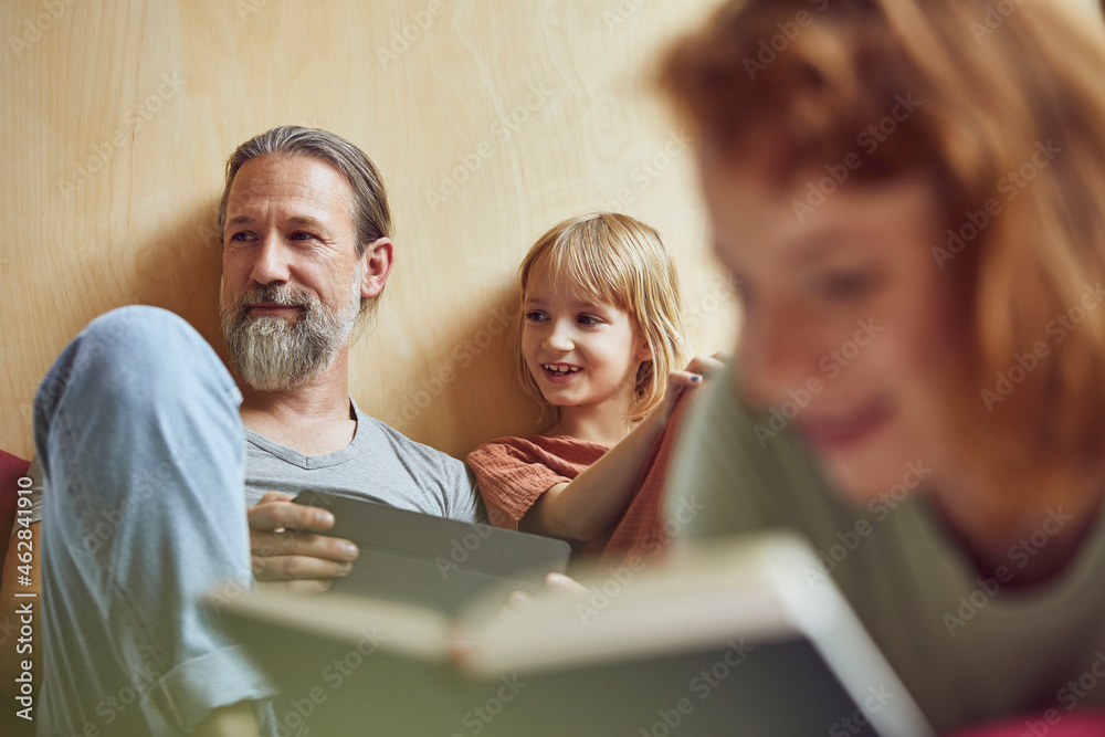 Woman reading book while father and daughter looking away in background at home Woman reading book while father and daughter looking away in background at home