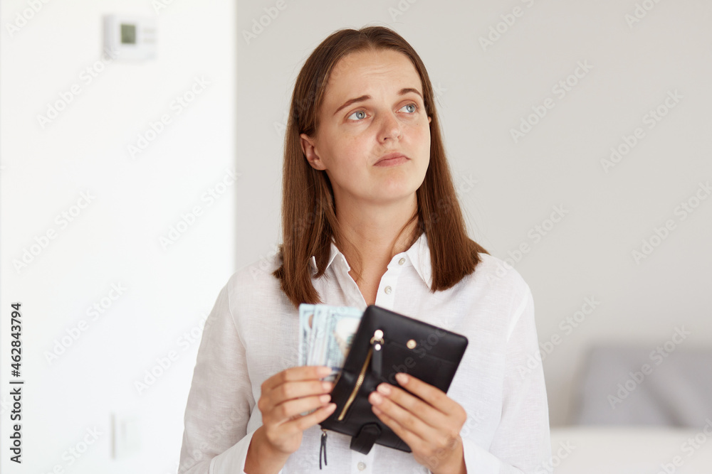 Indoor shot of sad upset dark haired female wearing white casual style shirt, holding wallet with money in hands and looking away, expressing sadness, have not enough money for shopping. Indoor shot of sad upset dark haired female wearing white casual style shirt, holding wallet with money in hands and looking away, expressing sadness, have not enough money for shopping.