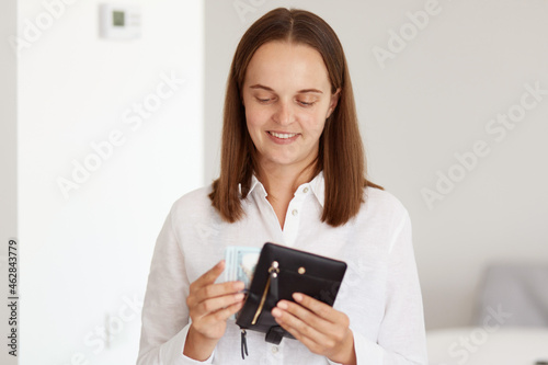 Portrait of happy dark haired female wearing white casual style shirt, holding wallet with banknotes in hands, counting her money, having positive facial expression.