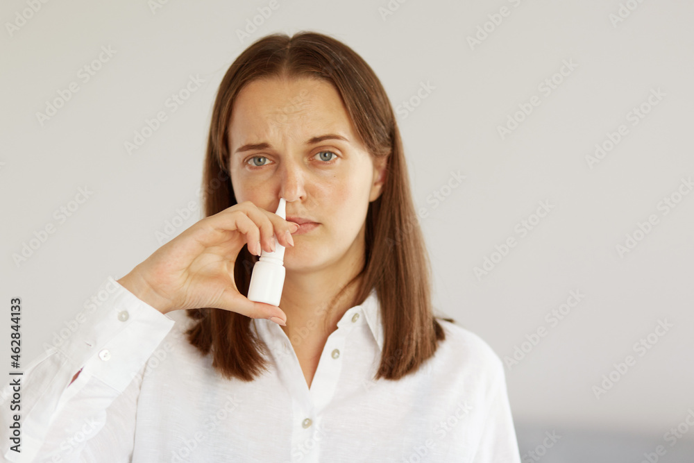 Indoor shot of sick winsome female with pleasant appearance wearing white casual style shirt, using nasal spray, suffering runny nose, looking at camera with ill frowning face. Indoor shot of sick winsome female with pleasant appearance wearing white casual style shirt, using nasal spray, suffering runny nose, looking at camera with ill frowning face.