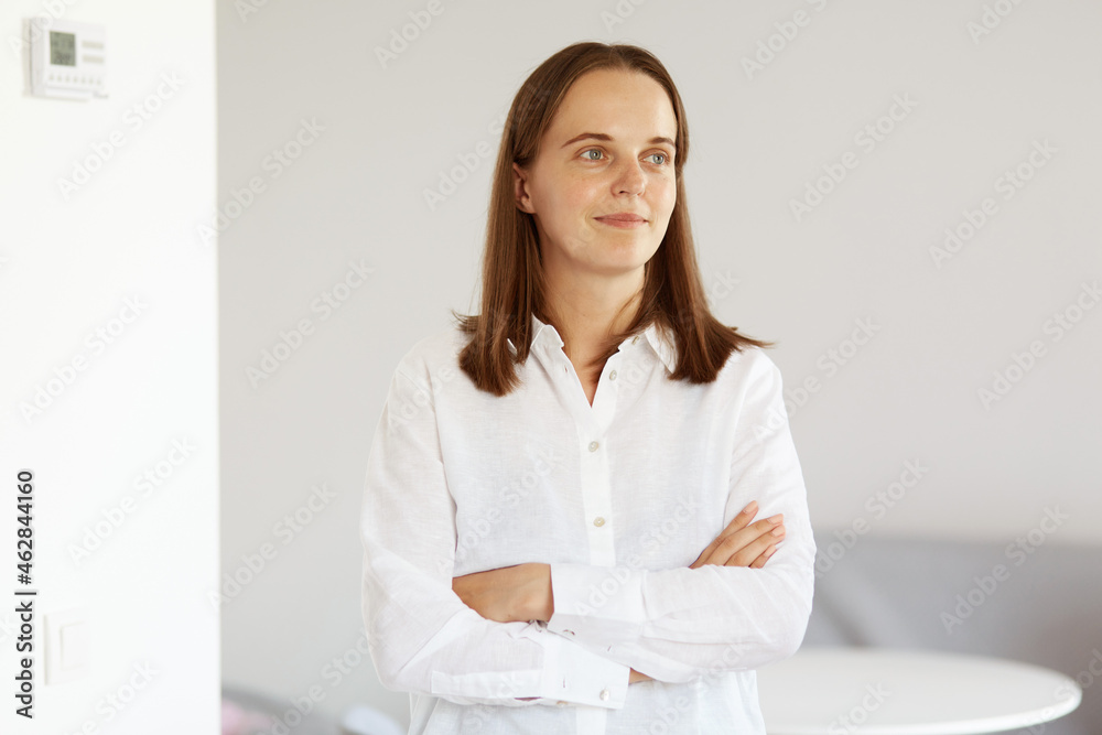 Portrait of attractive young adult woman with dark hair wearing white casual style shirt, standing with folded arms, looking away with confident expression. Portrait of attractive young adult woman with dark hair wearing white casual style shirt, standing with folded arms, looking away with confident expression.