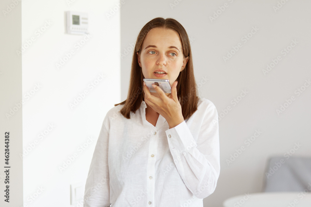 Young adult female with dark hair and pleasant appearance, wearing white casual style shirt, looking away, posing in light room at home, recording voice message or command. Young adult female with dark hair and pleasant appearance, wearing white casual style shirt, looking away, posing in light room at home, recording voice message or command.