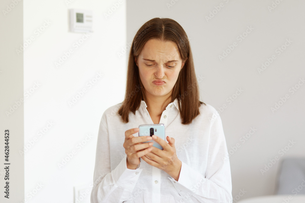 Indoor shot of sad upset female with dark hair wearing white casual style shirt, using mobile phone, reading bad news, expressing negative emotions, posing in light room at home. Indoor shot of sad upset female with dark hair wearing white casual style shirt, using mobile phone, reading bad news, expressing negative emotions, posing in light room at home.