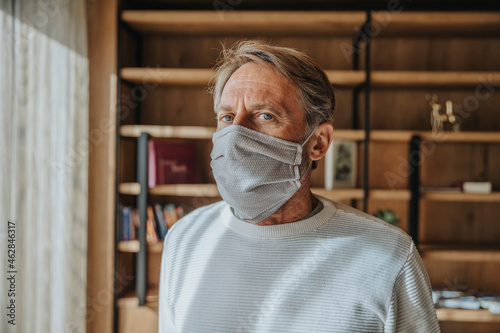 Mature man with gray eyes wearing protective face mask against shelf at home during COVID-19 photo