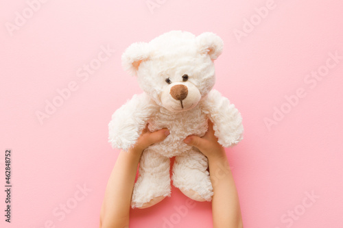 Smiling white teddy bear in baby girl hands on light pink background. Pastel color. Closeup. Point of view shot. Kids best friend. Top down view.