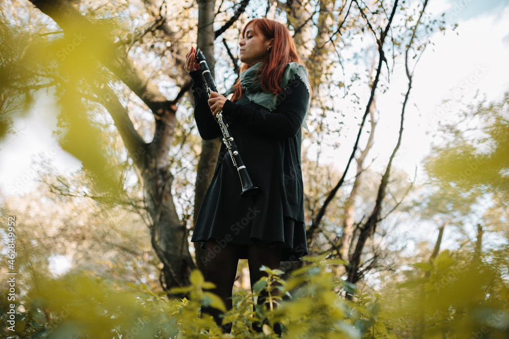 Redhead female musician with clarinet standing against bare trees in forest