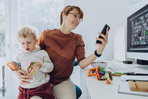 Mother using smart phone while son sitting on her lap playing with toys photo