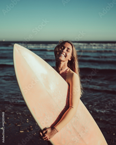 Cheerful woman with eyes closed carrying surfboard against sea during sunset photo