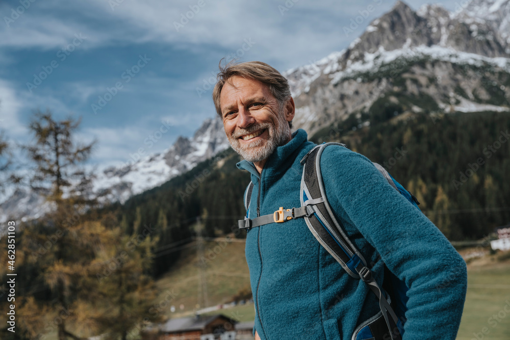Smiling mature man with backpack standing against sky at Hochkonig, Salzburger Land, Austria Smiling mature man with backpack standing against sky at Hochkonig, Salzburger Land, Austria