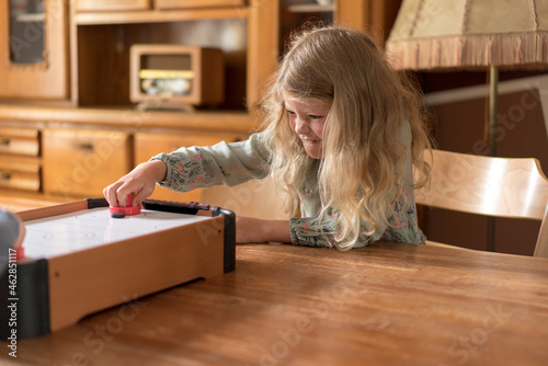 Blond girl playing air hockey while making faces on dining table at home photo