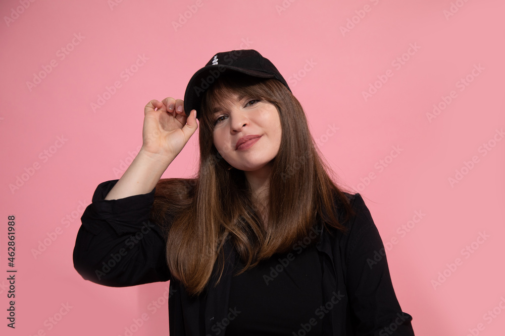 young dark-haired woman in baseball cap and black shirt holds cap with her hand by visor against pink backgroun, studio shot. Portrait. young dark-haired woman in baseball cap and black shirt holds cap with her hand by visor against pink backgroun, studio shot. Portrait.