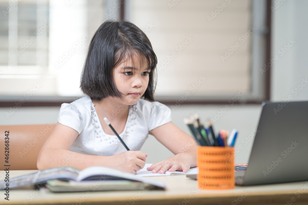 Asian children girl student writing on notebook while studying online via laptop while at home. Education Concept Stock Photo Asian children girl student writing on notebook while studying online via laptop while at home. Education Concept Stock Photo