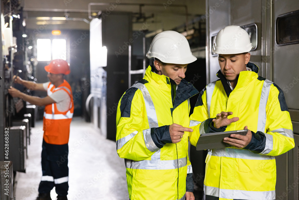 Male Electrical Engineers in Hard Hats Discuss New Project while Using Tablet Computer. They're Making Calculated Engineering Decisions. They Work at the Electric Power Factory Male Electrical Engineers in Hard Hats Discuss New Project while Using Tablet Computer. They're Making Calculated Engineering Decisions. They Work at the Electric Power Factory