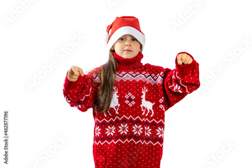 portrait of six-year-old girl in red Santa Claus hat in red knitted Christmas sweater with reindeer pointing fingers at camera, isolated on white background. 