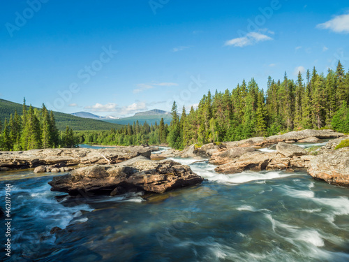 Beautiful landscape with long exposure water stream and cascade of river Kamajokk, boulders and spruce tree forest in Kvikkjokk village in Swedish Lapland. Summer sunny day, blue sky, white clouds