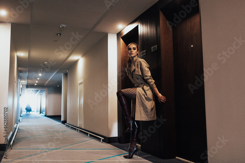 young beautiful girl in the hotel corridor, wearing a long trench coat and high boots
