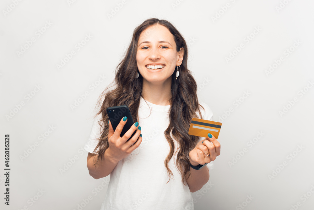 Pleased young girl is smiling at the camera while using phone and credit card she is holding. Pleased young girl is smiling at the camera while using phone and credit card she is holding.