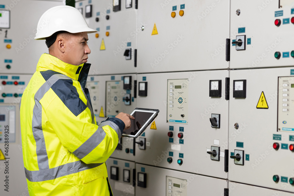 Asian man electrical engineer hold tablet monitoring electrical system in control room. Technician inspect to control panel screen system for generate electricity of factory in manufacture industrial Asian man electrical engineer hold tablet monitoring electrical system in control room. Technician inspect to control panel screen system for generate electricity of factory in manufacture industrial