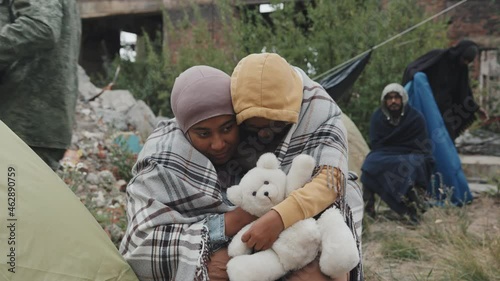 Medium shot of African-American mother and her 8-year-old daughter in warm clothing sitting outdoors under blanket embracing living at refugee camp with other immigrants photo