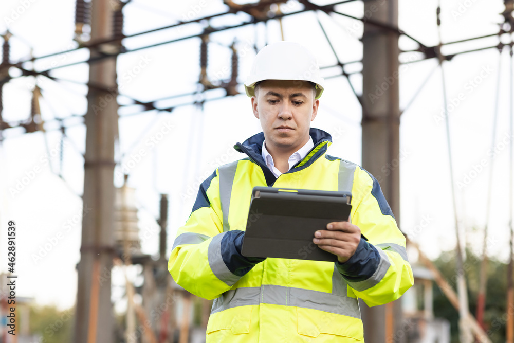 An energy engineer man in special clothes inspects a power line using data from electric sensors on a digital tablet computer. Electrical engineer with high voltage electricity pylon An energy engineer man in special clothes inspects a power line using data from electric sensors on a digital tablet computer. Electrical engineer with high voltage electricity pylon
